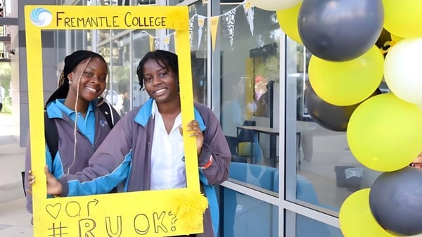 Dos estudiantes están frente a un cartel del Fremantle College con globos de colores en el fondo.