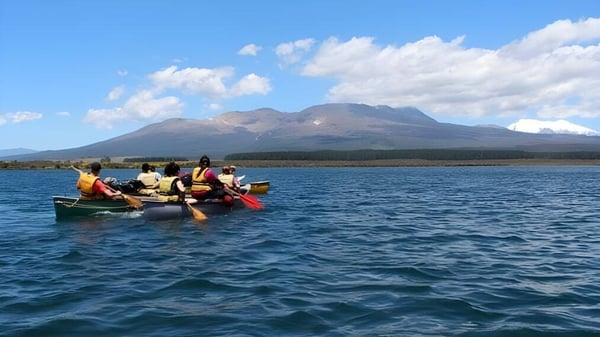 Estudiantes de la Freyberg High School reman en un pequeño bote en un lago frente a un paisaje montañoso.