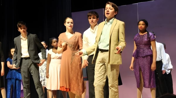 Un grupo de estudiantes de la Friends School of Baltimore está vestido formalmente en un escenario con un fondo violeta.