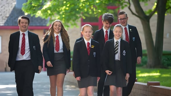 Un grupo de estudiantes en uniformes escolares camina juntos por el campus de Fulston Manor con árboles y edificios de fondo.