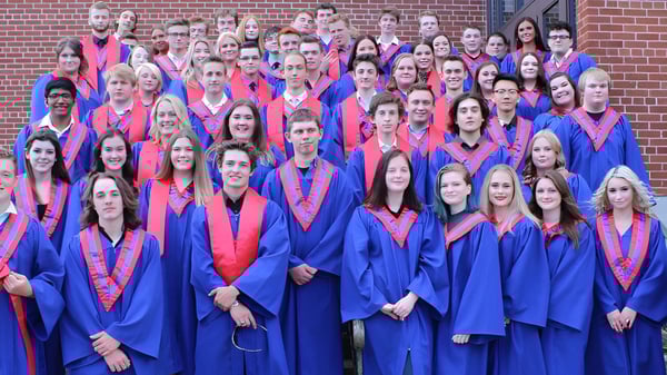 Un gran grupo de graduados de la Gananoque Secondary School está de pie en togas azules y rojas frente a un edificio de ladrillo.