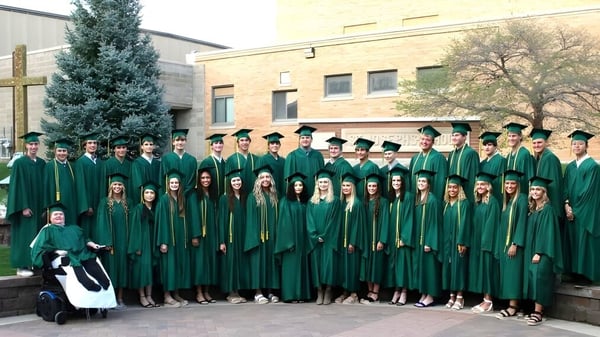 Los graduados de la Gehlen Catholic School están de pie en togas verdes frente a un edificio con un alto árbol perenne al fondo.