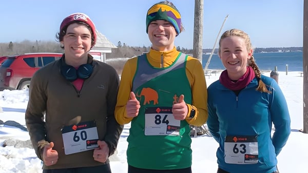 Tres personas con ropa de invierno posan frente a un paisaje nevado en el campus de la George Stevens Academy.