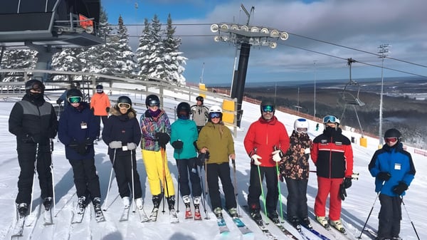 Un grupo de estudiantes de la Georgian Bay District Secondary School están en colorido equipo de esquí en una pendiente nevada con un telesilla y montañas de fondo.