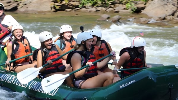 Alumnas de la Girls Preparatory School están sentadas con chalecos salvavidas y cascos en una balsa verde en un río con orillas rocosas y densa vegetación.