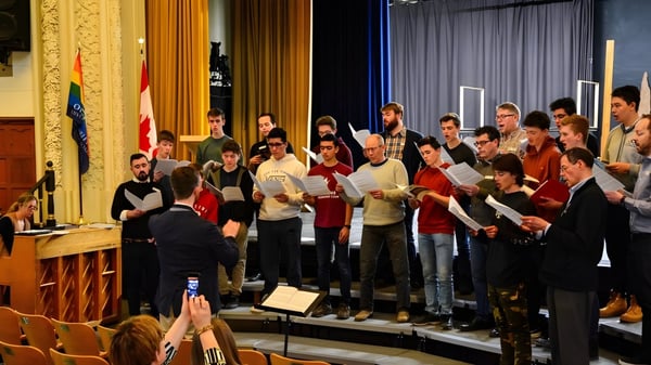 Un coro del Glebe Collegiate Institute se presenta en un escenario con elementos decorativos y banderas de fondo.