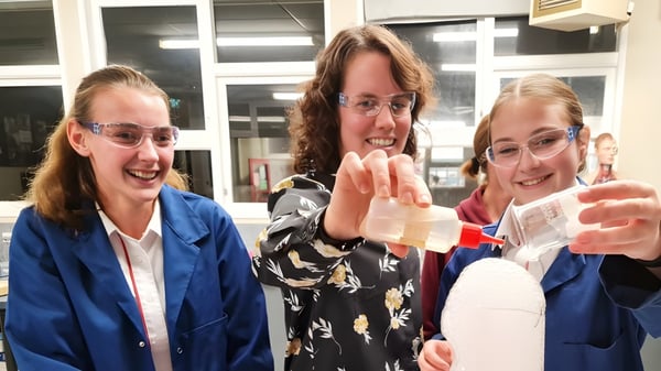 Tres estudiantes del Glenfield College están sonriendo y gesticulando en una habitación con ventanas y estanterías.