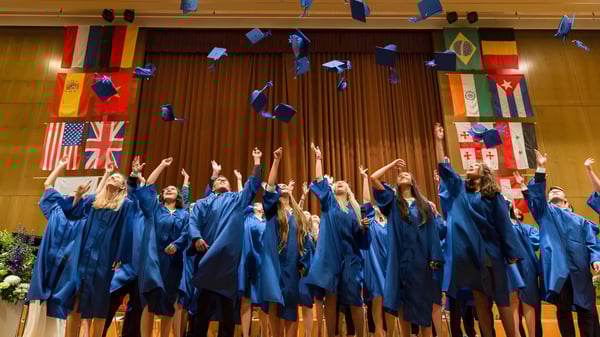 Las graduadas y graduados de la Glenlyon Norfolk School celebran con togas azules en un escenario con banderas internacionales.
