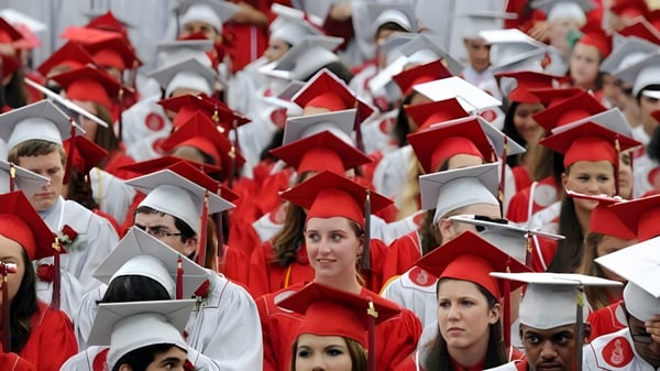 Un gran grupo de estudiantes de Gloucester High School lleva togas y birretes rojos frente a un fondo neutral.