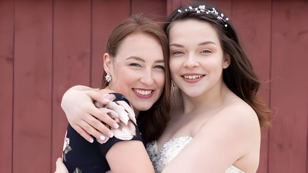 Dos mujeres sonrientes en ropa formal están frente a una pared de madera roja en el terreno de la Glovertown Academy.