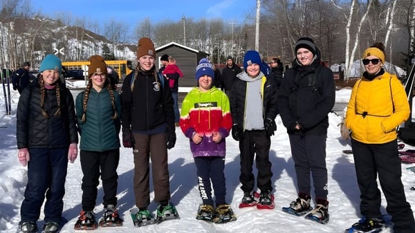 Un grupo de estudiantes de la Glovertown Academy está en invierno con ropa abrigada frente a un edificio de madera en la nieve.
