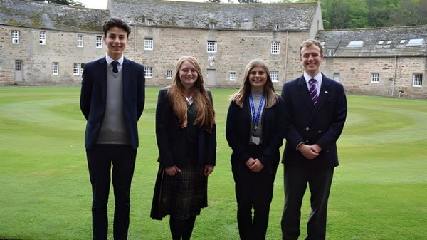 Cuatro personas están juntas en el patio cubierto de hierba de la Gordonstoun School entre edificios de piedra.