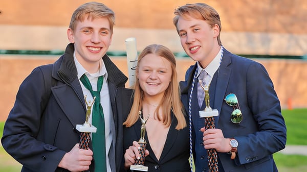 Tres estudiantes de la Grand Rapids Christian High School están afuera frente a una cerca de madera y árboles juntos.