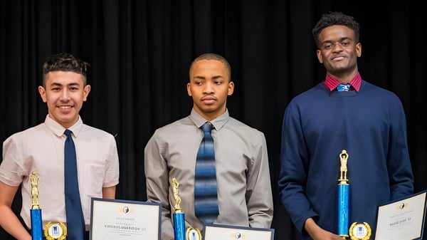Tres jóvenes en ropa formal sostienen certificados frente a un telón oscuro en la Grand River Academy.