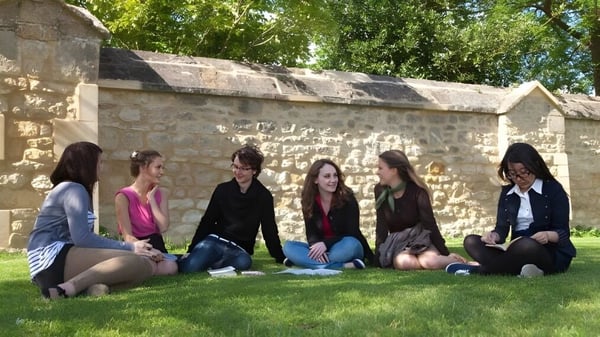 Un grupo de estudiantes se sienta en el campo frente a un muro de piedra en el campus de Greene's College Oxford.