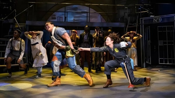 Un grupo de personas está en el escenario con un gran fondo de ventanales arqueados en el campus de la Groton School.