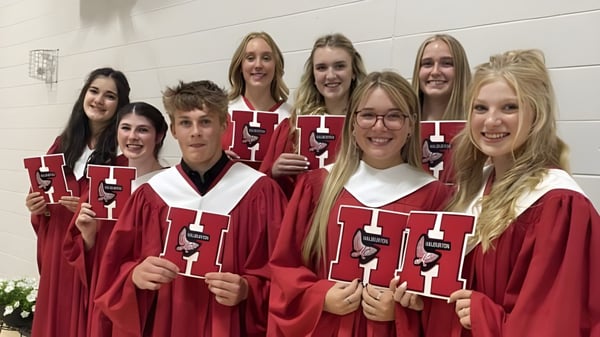 Un grupo de graduados en togas rojas sostiene sus diplomas frente a una pared blanca en el campus de la Haliburton Highlands School.