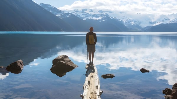 Un solo estudiante está en un muelle de madera frente a un lago espejo con montañas nevadas al fondo en el terreno de la Harbour View High School.