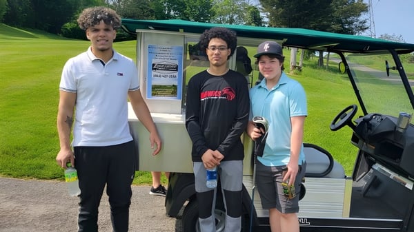 Tres estudiantes de la Harmony Christian School están frente a un carro de golf en un campo de hierba con árboles al fondo.