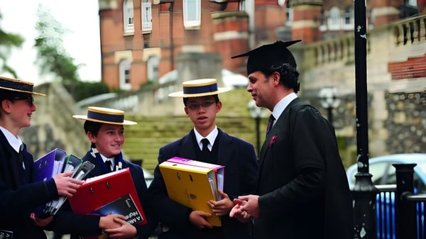 Un grupo de estudiantes de la Harrow School en ropa de graduación está en una calle frente a un edificio de ladrillo sosteniendo libros y documentos.