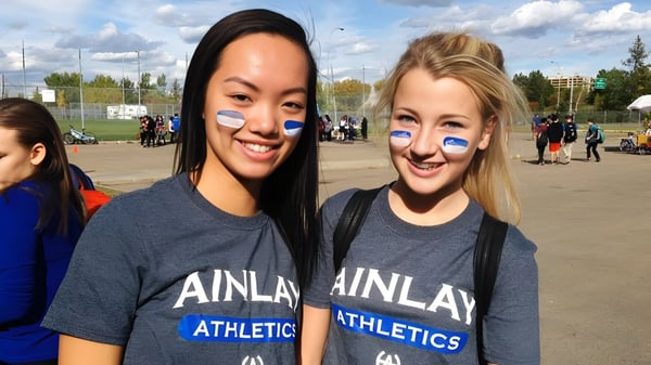 Dos estudiantes de la Harry Ainlay High School están sonriendo frente a un campo deportivo con otras personas al fondo.