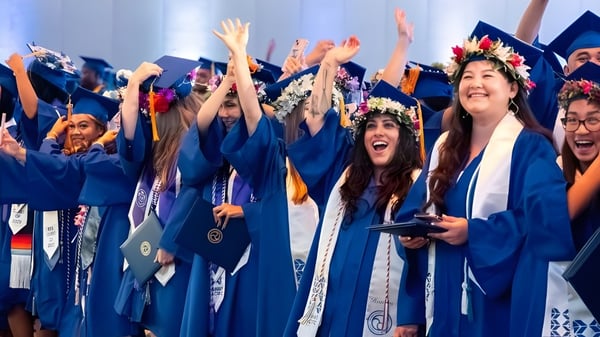 Un grupo de graduadas y graduados de la Hawai'i Pacific University celebra con los brazos en alto en togas azules y con coronas de flores.