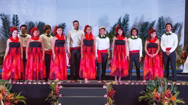 Estudiantes de la Hawaiian Mission Academy están en disfraces rojos y blancos en un escenario con un fondo tropical.