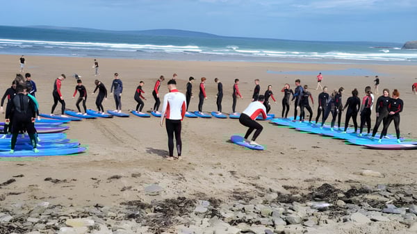 Un grupo de estudiantes del Hazelwood College está en trajes de neopreno en la playa con tablas de surf frente al océano.