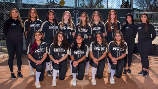 Un grupo de jóvenes deportistas de la Heritage Christian School posan juntas en el campo deportivo frente a una cerca y árboles.