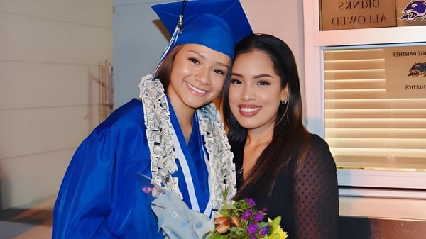 Dos estudiantes de la Heritage High School están juntas en sus togas de graduación en una habitación con ventana y decoración.