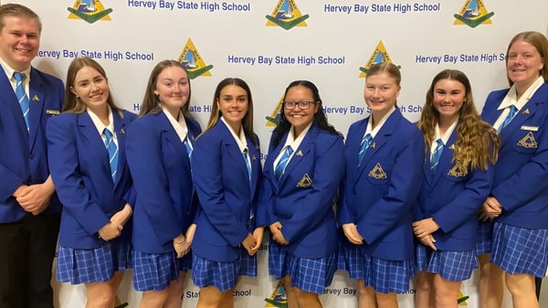 Un grupo de estudiantes de la Hervey Bay State High School está frente a un fondo con el nombre y el logo de la escuela.