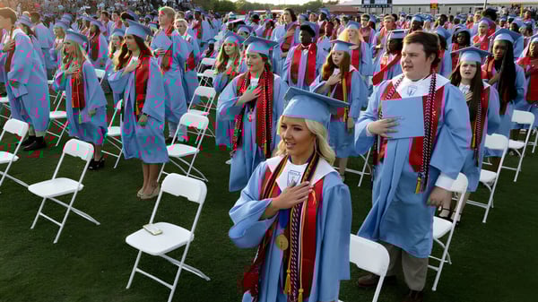 Un gran grupo de graduados está sentado en el césped durante la ceremonia de graduación de la Hillcrest High School.