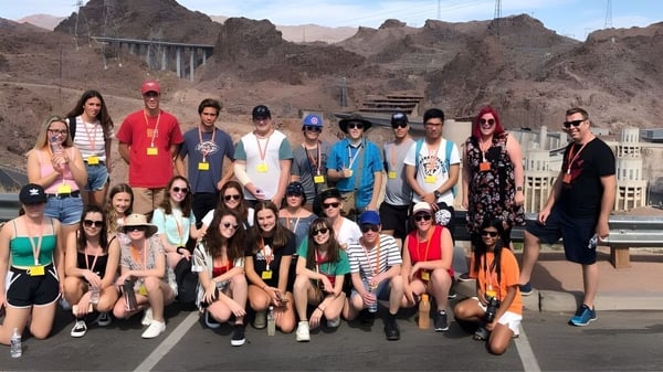 Un grupo de estudiantes de la Hillcrest High School posa frente a un dramático paisaje montañoso con un puente al fondo.