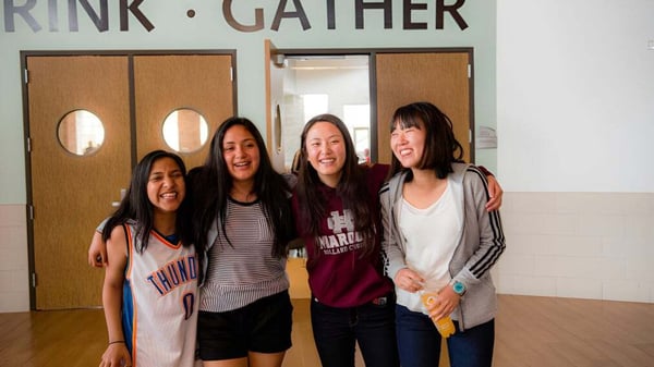 Cuatro estudiantes posan sonrientes juntas en una sala con un cartel de Rink Gather en la pared de Holland Christian Schools.