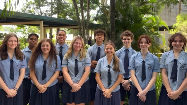 Un grupo de estudiantes de la Holland Park State High School está reunido en un área exterior verde con árboles y edificios escolares al fondo.