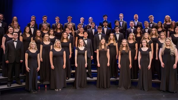Un grupo de alumnas y alumnos de la Holy Heart of Mary High School está de pie con ropa formal en un escenario con fondo azul.