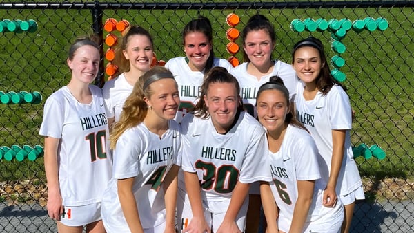 Un grupo de jóvenes deportistas en uniformes blancos posan frente a una cerca verde en el campus de la Hopkinton High School.