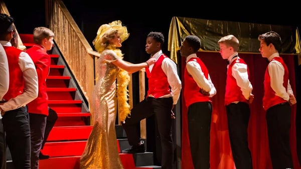 Estudiantes de Houghton Academy llevan togas rojas en la fiesta de graduación en un escenario con cortina dorada.