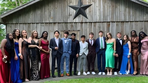Un grupo de personas en ropa formal está frente a un establo rústico con un gran emblema estrellado en el campus de la Houghton Academy.