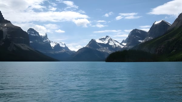 Un tranquilo lago de montaña está rodeado de montañas cubiertas de nieve bajo un cielo parcialmente nublado.