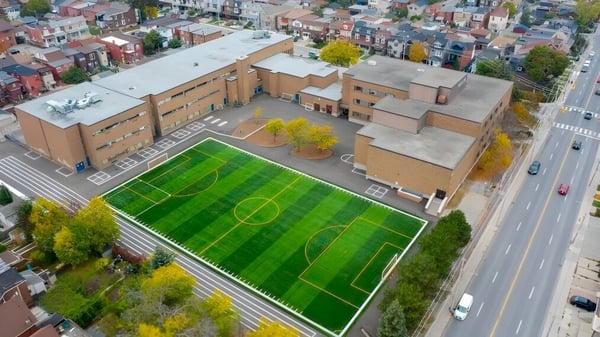 El gran campo de fútbol verde en el campus del Hudson College con edificios y una calle concurrida de fondo.