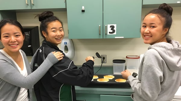 Tres estudiantes interactúan sonriendo en una cocina en el campus de la Hugh McRoberts Secondary School.