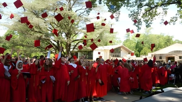 Un grupo de estudiantes de The Hun School of Princeton está de pie en togas de graduación rojas bajo árboles con decoraciones rojas.
