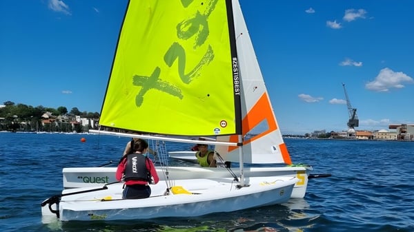 Un pequeño velero con vela amarillo-naranja navega en el agua frente a edificios cerca de la Hunters Hill High School.