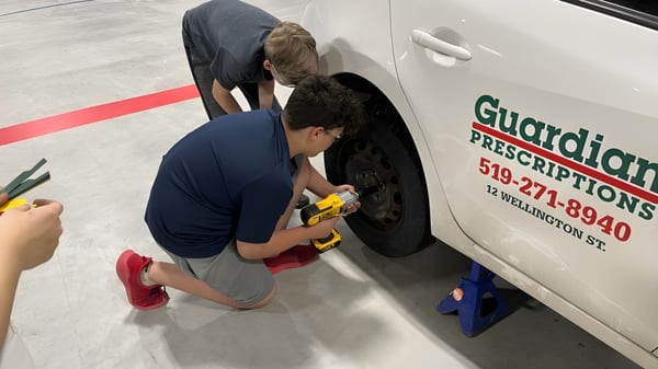 Una persona está de rodillas en el suelo junto a un furgón del Huron-Perth Catholic District School Board e inspecciona el vehículo.