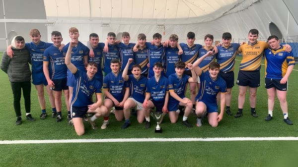 Un grupo de jóvenes atletas del Iascaigh College posan en un campo deportivo frente a un gran gimnasio.