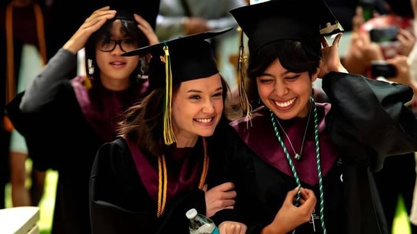 Dos graduadas de la Idyllwild Arts Academy se abrazan en sus togas de graduación frente a un grupo de otros graduados.