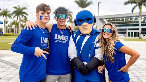 Un grupo de cuatro estudiantes lleva uniformes azules de la IMG Academy frente a un edificio con palmeras.