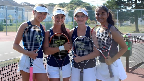 Cuatro estudiantes de la Immaculata High School están con raquetas de tenis en una cancha de tenis rodeada de árboles y una cerca.