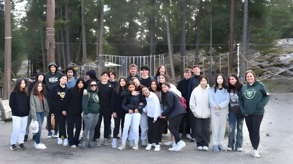 Un grupo de alumnas y alumnos de la Immaculata Regional School está junto en un área boscosa con un puente de cuerdas al fondo.